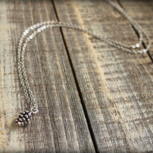 May include: A silver chain necklace with a small, detailed pine cone pendant. The necklace is lying on a rustic wooden surface.