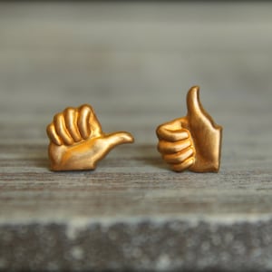 May include: Two gold-coloured thumbs up earrings. One thumb is pointing to the side, the other is pointing upwards.
