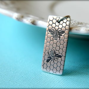 May include: Silver pendant necklace with a honeycomb design and two bees. The pendant is rectangular and has a textured surface.