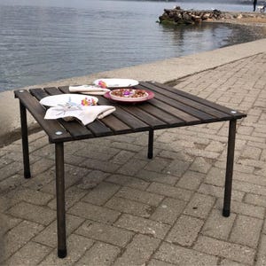 Puede incluir: Una mesa cuadrada de madera con listones marrones oscuros y patas de metal. Dos platos blancos, un plato rojo con comida y dos servilletas blancas dobladas con diseños florales están sobre la mesa. La mesa está puesta al aire libre cerca de un cuerpo de agua.