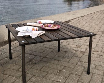 May include: A square wooden table with dark brown slats and metal legs. Two white plates, a red plate with food, and two folded white napkins with floral designs are on the table. The table is set outdoors near a body of water.