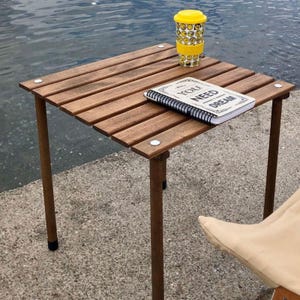 Può includere: Un tavolino quadrato in legno con gambe marroni e piano a doghe. Una tazza da viaggio gialla con un design a faccine sorridenti e un quaderno a spirale con la scritta "ALL YOU NEED IS DREAM" sono appoggiati sopra. Il tavolo misura circa 61 cm quadrati.