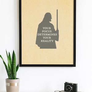 Puede incluir: Impresión enmarcada con la silueta de una figura sosteniendo un bastón, con el texto "YOUR FOCUS DETERMINES YOUR REALITY". La obra de arte enmarcada en negro está sobre un escritorio blanco, con una planta y una cámara.