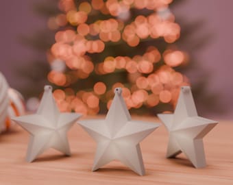 Puede incluir: Tres adornos navideños blancos en forma de estrella con un brillo suave, apoyados sobre una superficie de madera. Los adornos tienen un diseño simple y moderno. Un árbol de Navidad borroso con luces cálidas en el fondo.
