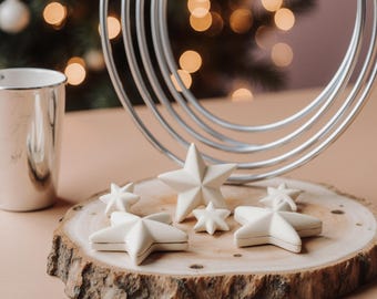 Puede incluir: Una colección de adornos blancos en forma de estrella de varios tamaños dispuestos sobre una rodaja de madera. Un anillo de metal plateado y una taza plateada están en el fondo. La escena se desarrolla contra un fondo borroso de un árbol de Navidad con luces cálidas y brillantes.
