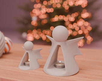 May include: Two white angel candle holders with lit tea lights. The angel figures have a minimalist design with a round head and outstretched wings. The background features a blurred Christmas tree with warm, glowing lights, creating a festive atmosphere.