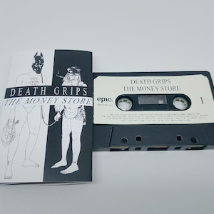 Könnte beinhalten: Eine schwarze Kassette und ein Booklet mit dem Text "DEATH GRIPS THE MONEY STORE". Die Kassette hat ein weißes Etikett mit schwarzem Text. Das Booklet zeigt Schwarz-Weiß-Illustrationen von Figuren.