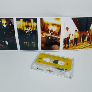 May include: A clear cassette tape with a yellow label and the number 2 is in the foreground. Behind it is a folded album cover with several images of people in various settings, with a warm color palette.