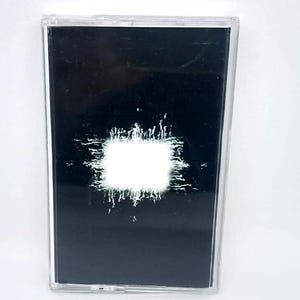 May include: A black cassette tape with a white, textured square design in the center. The cassette is in a clear plastic case. The design has a grunge aesthetic.