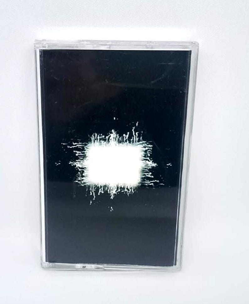 May include: A black cassette tape with a white, textured square design in the center. The cassette is in a clear plastic case. The design has a grunge aesthetic.