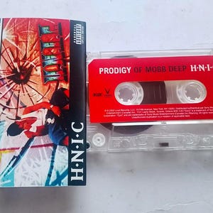 May include: A vintage cassette tape of Prodigy of Mobb Deep's album, H.N.I.C. The red cassette is in a clear plastic case, with the album title and artist's name printed on the tape. The album cover is visible, featuring artwork with the album title.