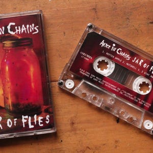 Könnte beinhalten: Eine Kassette und ihre Hülle für das Album "Jar of Flies" von Alice in Chains. Die Hülle zeigt ein rot getöntes Bild eines Glases und eines Gesichts. Die Kassette ist transparent mit einem roten Etikett, das den Albumtitel und die Titelliste anzeigt.