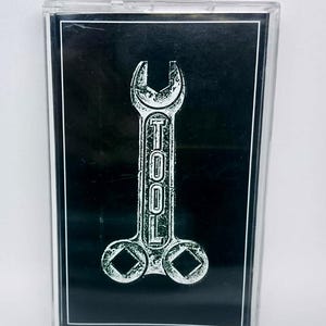 May include: A cassette tape with a black background and a white graphic of a wrench with the word "TOOL" printed vertically. The cassette case is clear.