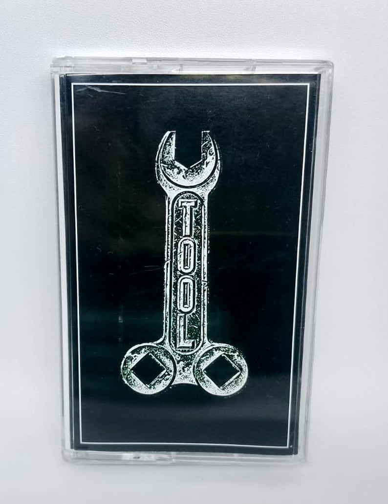 May include: A cassette tape with a black background and a white graphic of a wrench with the word "TOOL" printed vertically. The cassette case is clear.