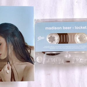 Może przedstawiać: Kaseta magnetofonowa i okładka albumu z piosenkarką Madison Beer. Kaseta jest przezroczysta z jasnoniebieską etykietą z napisem "madison beer - locket". Okładka albumu przedstawia portret Madison Beer.