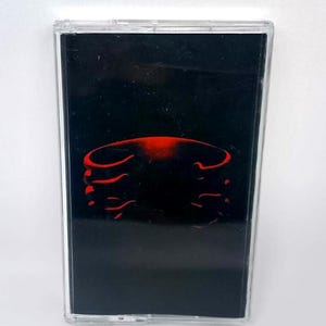 May include: A cassette tape with a black background and a red abstract design. The cassette is in a clear plastic case. The design appears to be a stylized rib cage.