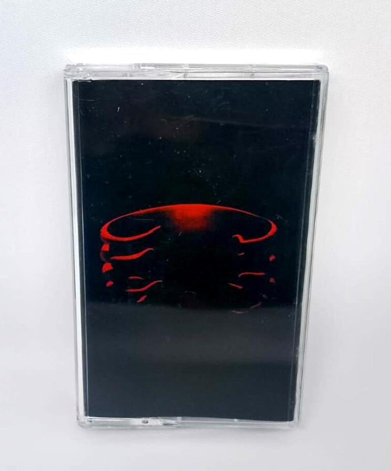 May include: A cassette tape with a black background and a red abstract design. The cassette is in a clear plastic case. The design appears to be a stylized rib cage.