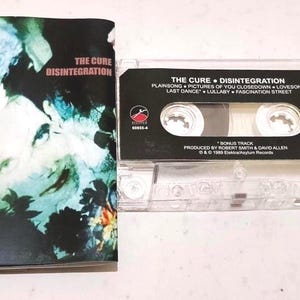 Könnte beinhalten: Eine Musikkassette und das Albumcover für The Cure's Album Disintegration. Das Albumcover zeigt ein surreales Bild mit einem Gesicht und floralen Elementen. Die Kassette ist transparent mit einem schwarzen Etikett, das den Albumtitel und die Titelliste anzeigt.