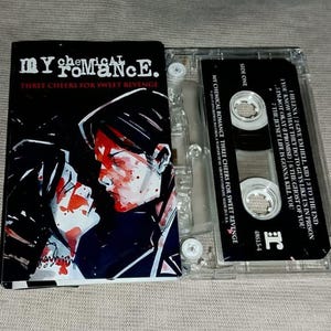 May include: A cassette tape of the album "Three Cheers for Sweet Revenge" by My Chemical Romance. The album cover features a black and white illustration of two figures with red accents. The cassette case is clear.