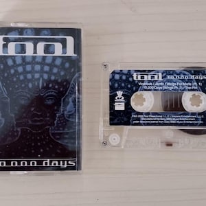 Può includere: Una musicassetta trasparente con la copertina dell'album "10,000 Days" dei Tool. La copertina presenta un design blu scuro e nero con il logo della band e il titolo dell'album. La cassetta è accanto alla sua custodia.