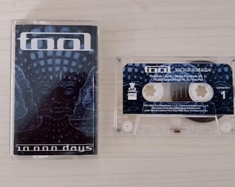 May include: A clear cassette tape with the album art for Tool's "10,000 Days" album. The cover art features a dark blue and black design with the band's logo and album title. The cassette is next to its case.