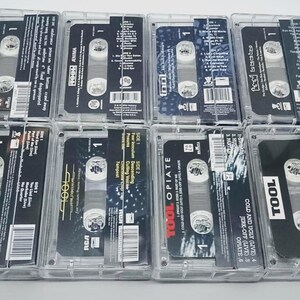 May include: A collection of eight cassette tapes in clear plastic cases. The tapes feature black and white labels with the band name "TOOL" and album titles. The cases are arranged on a white surface.