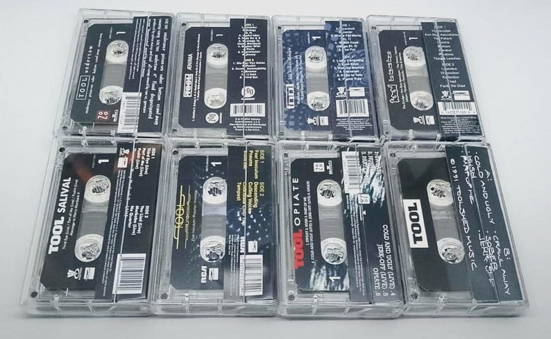 May include: A collection of eight cassette tapes in clear plastic cases. The tapes feature black and white labels with the band name "TOOL" and album titles. The cases are arranged on a white surface.