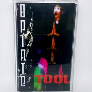 May include: A cassette tape with a black and dark background. The word "Opiate" is printed vertically in white on the left side. The word "TOOL" is printed in red at the bottom. The cover art features a figure with hands clasped in prayer.