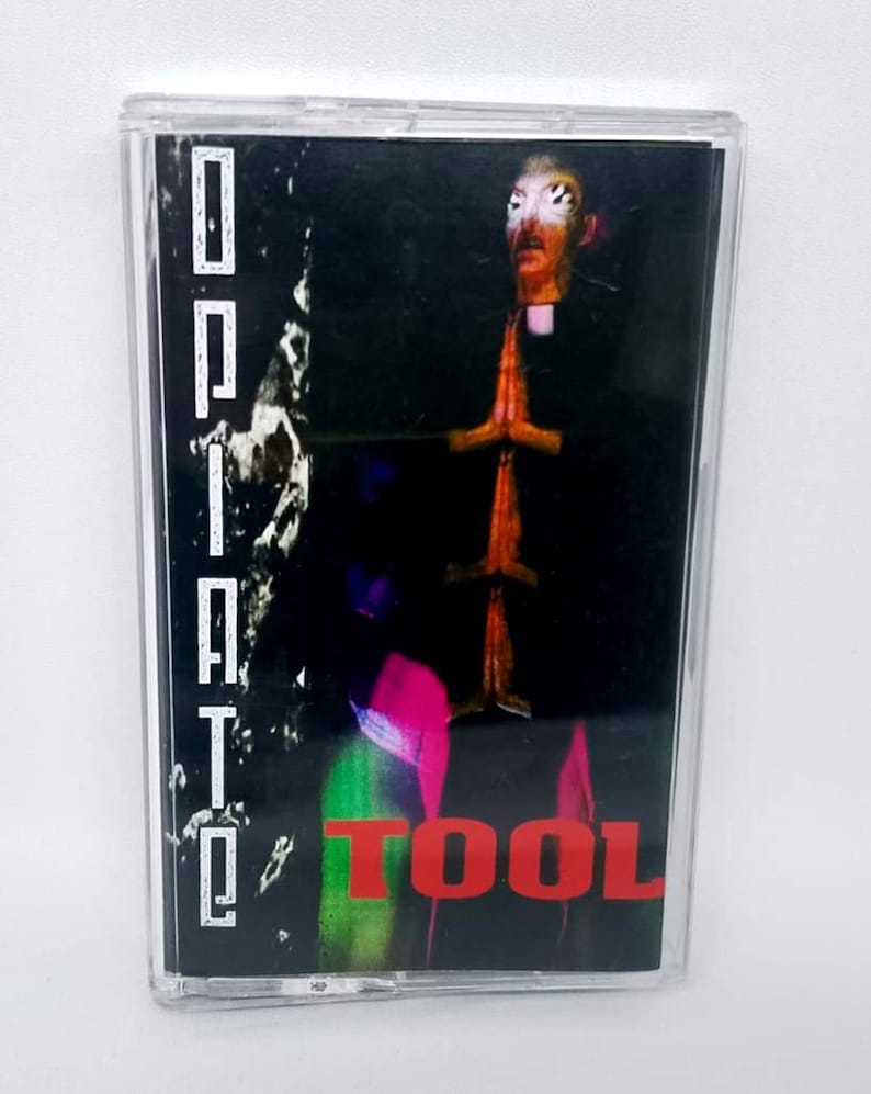 May include: A cassette tape with a black and dark background. The word "Opiate" is printed vertically in white on the left side. The word "TOOL" is printed in red at the bottom. The cover art features a figure with hands clasped in prayer.