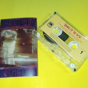 May include: A cassette tape and its case are shown on a yellow surface. The cassette case has a blurry image with purple text that reads "Temple of the Dog". The cassette tape is clear with a yellow label that reads "Temple of the Dog".
