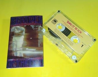 May include: A cassette tape and its case are shown on a yellow surface. The cassette case has a blurry image with purple text that reads "Temple of the Dog". The cassette tape is clear with a yellow label that reads "Temple of the Dog".