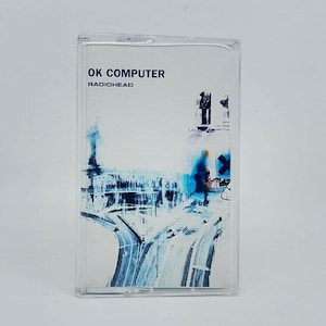 May include: A clear plastic cassette case containing the album OK Computer by Radiohead. The insert features a blue and white abstract graphic of a highway intersection and a blurred figure, with the text OK COMPUTER and RADIOHEAD printed in black.