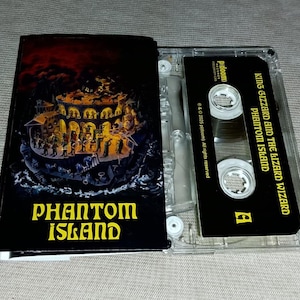 Könnte beinhalten: Eine Musikkassette des Albums "Phantom Island" von King Gizzard and the Lizard Wizard. Die Kassette ist geöffnet und zeigt das schwarze Band und das Cover. Das Cover zeigt eine detaillierte Illustration mit dem Albumtitel in Gelb.