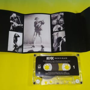 May include: A cassette tape of the AC/DC album "Back in Black" with a black and white photo insert. The cassette is clear with yellow accents and the album title is printed on the label. The insert features band photos.