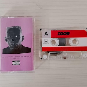 Puede incluir: Una cinta de casete del álbum "IGOR" de Tyler, The Creator. La caja del casete es transparente, con una portada rosa que presenta un retrato en blanco y negro. El casete en sí tiene una etiqueta roja y blanca con el título del álbum.