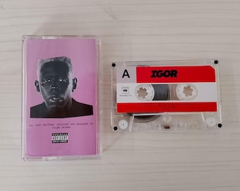 May include: A cassette tape of the album "IGOR" by Tyler, The Creator. The cassette case is clear, with a pink cover featuring a black and white portrait. The cassette itself has a red and white label with the album title.