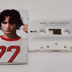 Op de afbeelding: Een witte cassetteband met de tekst "sombr / I Barely Know Her" en een foto van een persoon die een wit shirt met een rode kraag en het nummer "77" draagt. De cassette is gemarkeerd met het nummer "1".