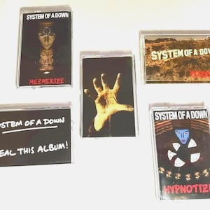 May include: A collection of five System of a Down cassette tapes. The tapes feature album art with text including "MEZMERIZE", "STEAL THIS ALBUM!", and "HYPNOTIZE". The artwork includes a hand, a face, and a landscape.