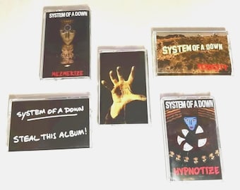 May include: A collection of five System of a Down cassette tapes. The tapes feature album art with text including "MEZMERIZE", "STEAL THIS ALBUM!", and "HYPNOTIZE". The artwork includes a hand, a face, and a landscape.