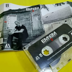 Puede incluir: Una cinta de cassette y la carátula del álbum de Eminem, The Marshall Mathers LP 2. La carátula presenta una imagen en blanco y negro de Eminem sentado en los escalones de una casa. La cinta de cassette es transparente con una etiqueta negra.