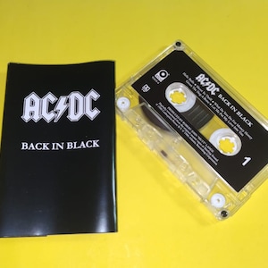 May include: A vintage AC/DC "Back in Black" cassette tape and its black case. The cassette is clear with a black label and white text. The case is black with the band's logo and album title in white.