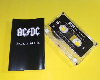 May include: A vintage AC/DC "Back in Black" cassette tape and its black case. The cassette is clear with a black label and white text. The case is black with the band's logo and album title in white.