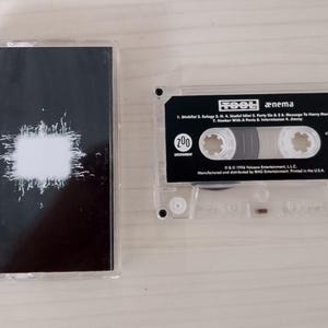 May include: A clear cassette tape with a black and white cover featuring a white blotch design. The cassette tape is labeled "TOOL ænima" with a tracklist and the number "1".