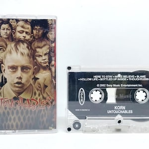 Puede incluir: Un casete de Korn Untouchables con caja de plástico transparente y carátula con un collage de rostros. La carcasa negra del casete muestra el título del álbum, la lista de canciones, el logo de Epic y el texto 2002 Sony Music Entertainment Inc. ET86696.
