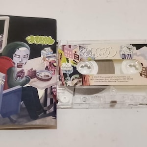 May include: A clear cassette tape and album art featuring the word "DOOM" in green. The album art depicts a figure in a mask seated at a table. The cassette tape has a label with the word "DOOM" and various graphics.