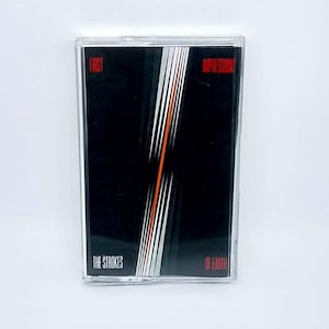 May include: A cassette tape with a black cover featuring white and orange diagonal lines. The text "FIRST IMPRESSIONS" and "OF EARTH" are in red, with "THE STROKES" in white.