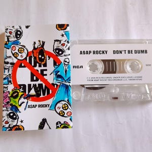Könnte beinhalten: Eine transparente Kassette und ihre Hülle. Die weiße Hülle zeigt bunte Cartoon-Illustrationen und den Text "DON'T BE DUMB" in einem roten Kreis. Auf der Kassette steht "ASAP ROCKY DON'T BE DUMB".