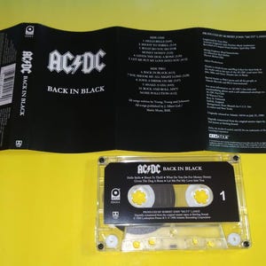 May include: AC/DC "Back in Black" cassette tape with the album cover and tracklist visible. The cassette is clear with black labels and the number 1. The album cover is black with white text.