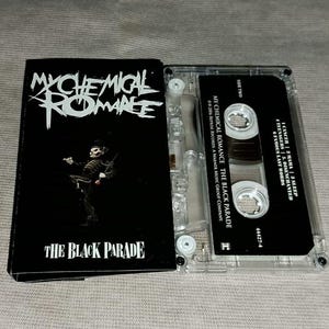 Pode incluir: Uma fita cassete preta e sua caixa com a banda My Chemical Romance. A caixa tem o logotipo da banda e o título do álbum "The Black Parade" em texto branco. A fita cassete é transparente com texto preto.