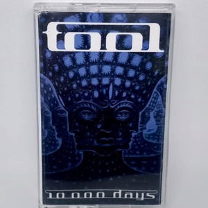 May include: A Tool cassette tape with a blue and black album cover. The cover features the band's logo in white at the top and the album title "10,000 Days" at the bottom. The artwork depicts multiple faces.